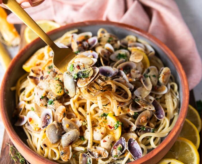 A fork stretching the clam pasta showing the details of the spaghetti and the cooked clams in the simple clam sauce with some lemon slices and chilli flakes