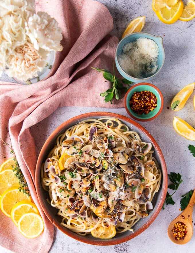 Plato de pasta de almeja completamente configurado rodeado de flores y algunas rodajas de limón, un paño de cocina, algunas especias y un tazón de queso parmesano fresco 