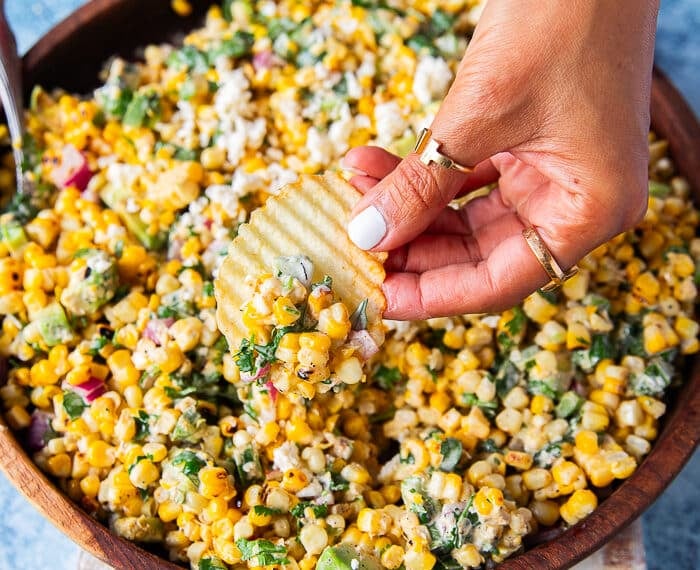 A hand holding a chips and scooping some mexican corn salad with creamy cilantro lime dressing, jalapenos and avocados