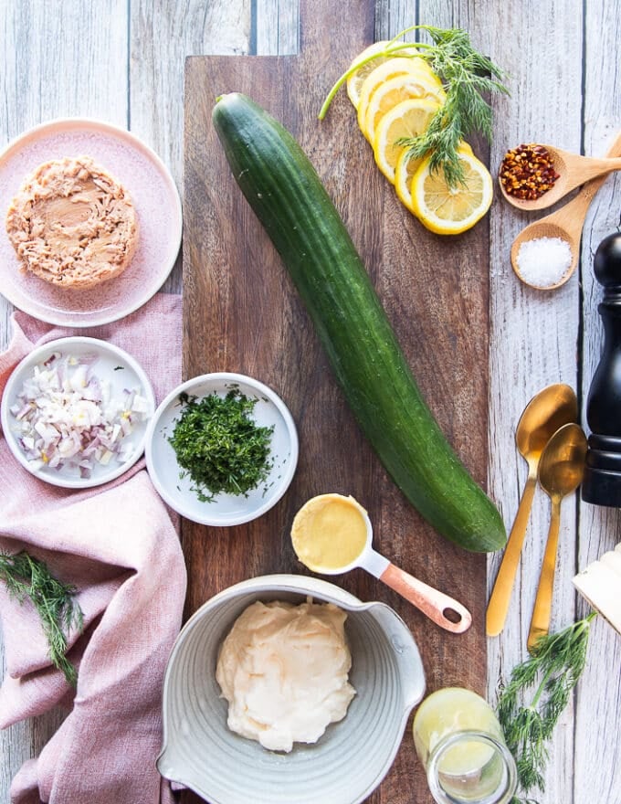 Ingredients for tuna cucumber salad including a whole cucumber, canned tuna in a bowl, fresh dill chopped, some mayo in a bowl, some spices, some onions chopped, lemon juice