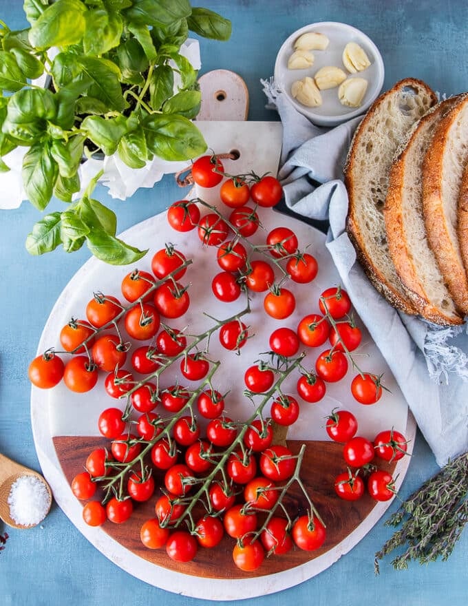 Ingredients to make caprese toast including baby tomatoes on the vine, some garlic, a jar of olive oil, some fresh basil leaves, some mozzarella cheese, sourdough toast , salt and pepper