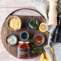 ingredients in small bowls t make harissa aioli including a bowl of mayo, harissa paste, a bowl of minced garlic, a bowl of minced cilantro, a bowl of lemon juice, some salt and pepper, a bowl of dried spice