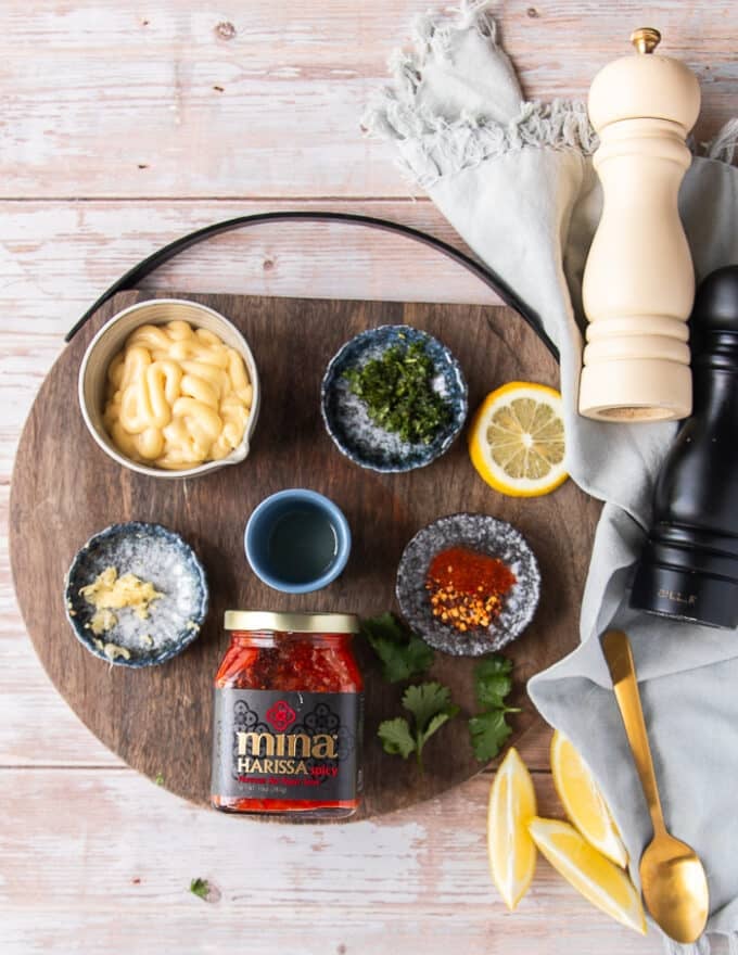 ingredients in small bowls t make harissa aioli including a bowl of mayo, harissa paste, a bowl of minced garlic, a bowl of minced cilantro, a bowl of lemon juice, some salt and pepper, a bowl of dried spice 