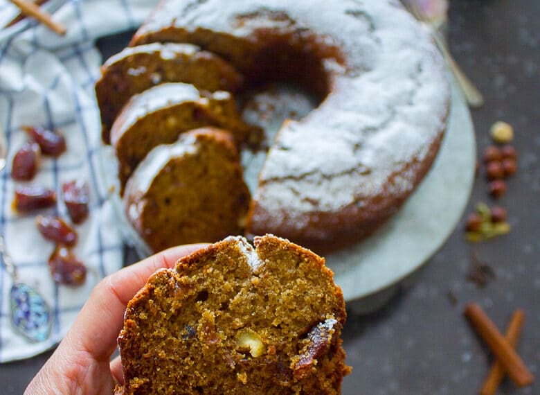 A hand holding a slice of date cake showing the smooth texture and chunky walnuts in the cake with bits of dates showing