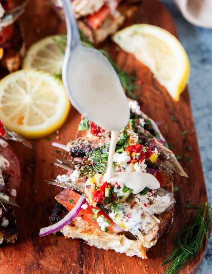 A spoon drizzling a hefty amount of tahini sauce over the sardines on toast 
