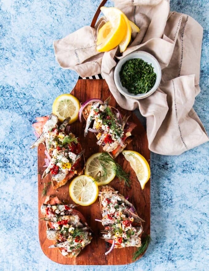 A board with fully dressed and ready to eat sardines on toast surrounded by fresh dill and lemon wedges for squeezing