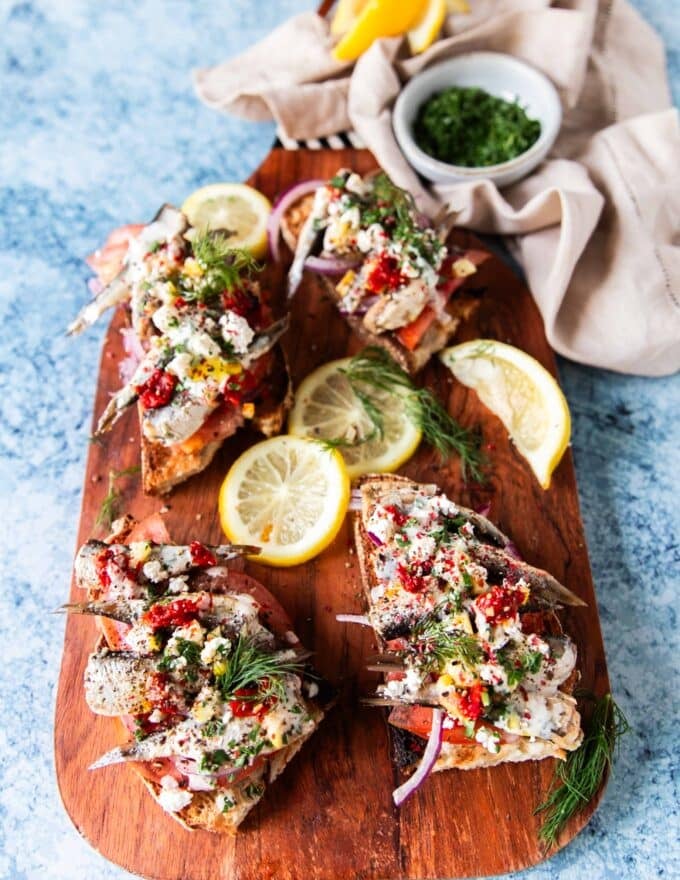a board with sardines on toasts topped with veggies and sauces, some lemon wedges on the side