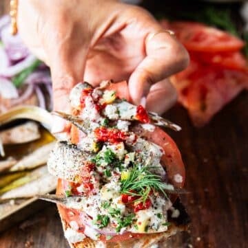 A hand holding a toast of sardines with all the toppings like lemon, feta onions, tomatoes, chilli flakes and more close up showing the texture and layering of the toast