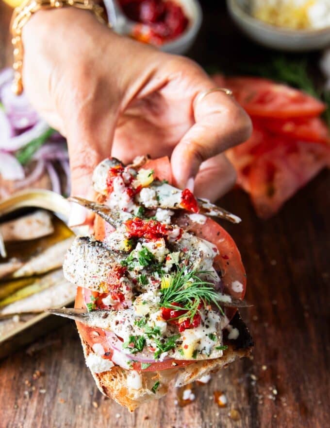 A hand holding a toast of sardines with all the toppings like lemon, feta onions, tomatoes, chilli flakes and more close up showing the texture and layering of the toast