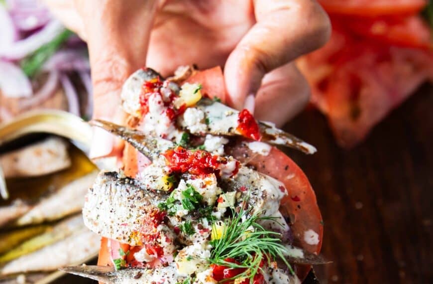 A hand holding a toast of sardines with all the toppings like lemon, feta onions, tomatoes, chilli flakes and more close up showing the texture and layering of the toast