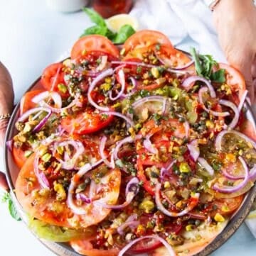 A hand holding a layered tomato carpaccio on a wooden bowl