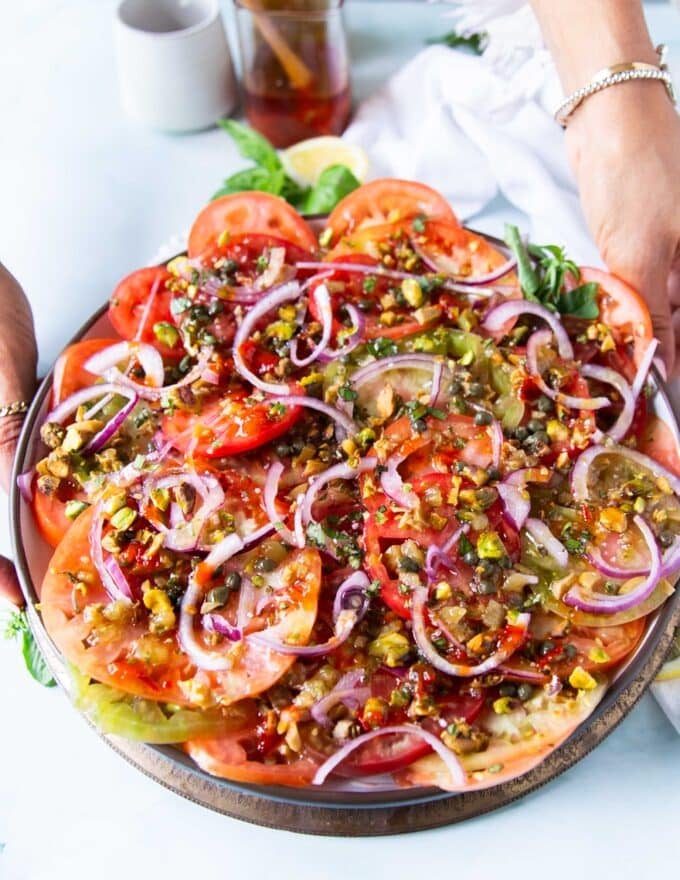 A hand holding a layered tomato carpaccio on a wooden bowl