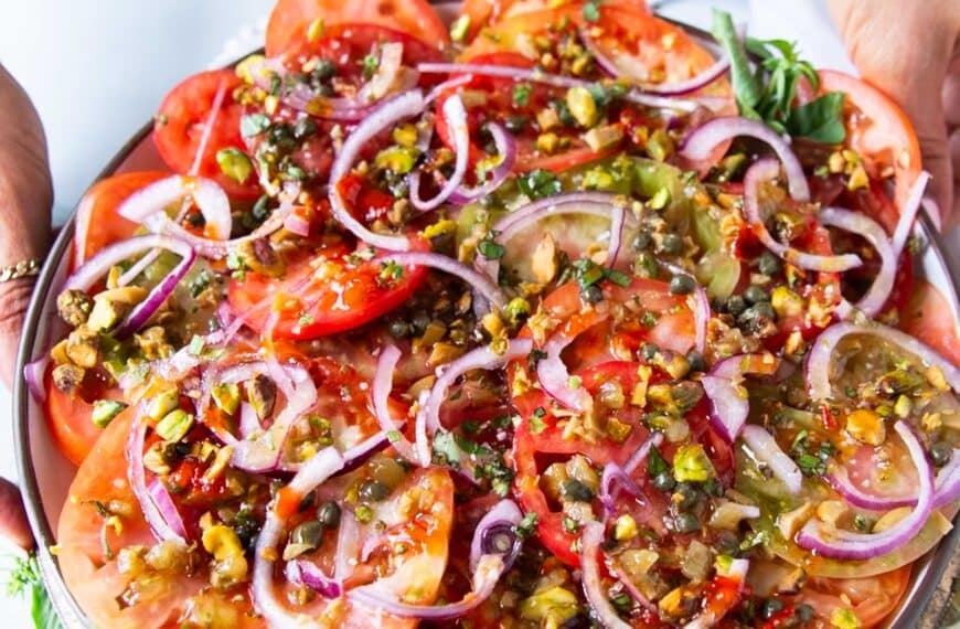 A hand holding a layered tomato carpaccio on a wooden bowl