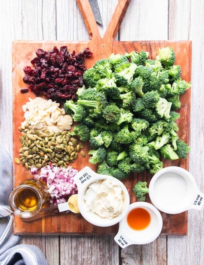 broccoli salad ingredients on a board with some fresh broccoli florets, dried cranberries, seeds, nuts and dressing ingredients