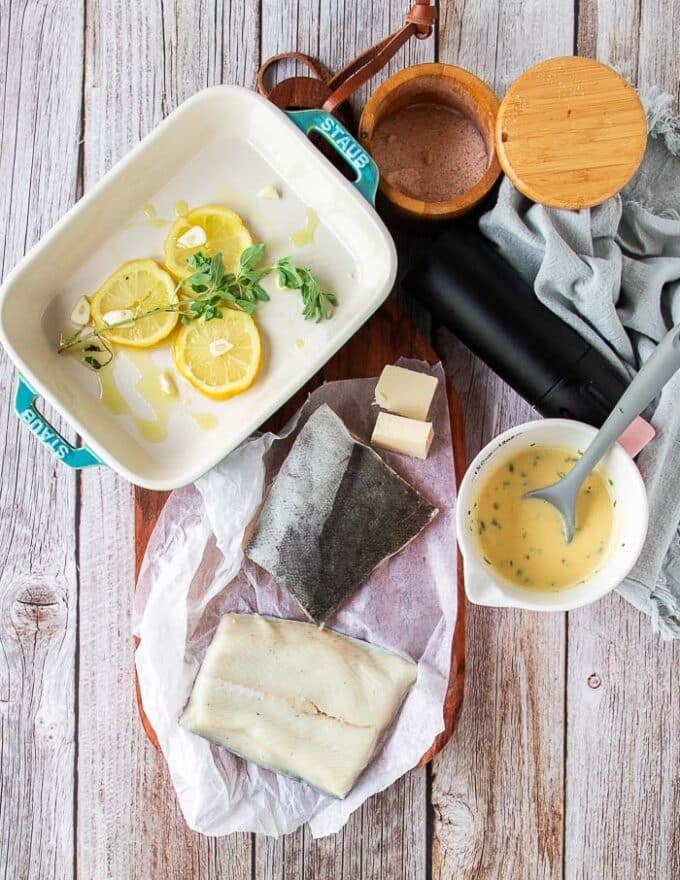 Ingredients for baked chilean sea bass on a board including the fish, butter, lemon, herbs and beurre blanc
