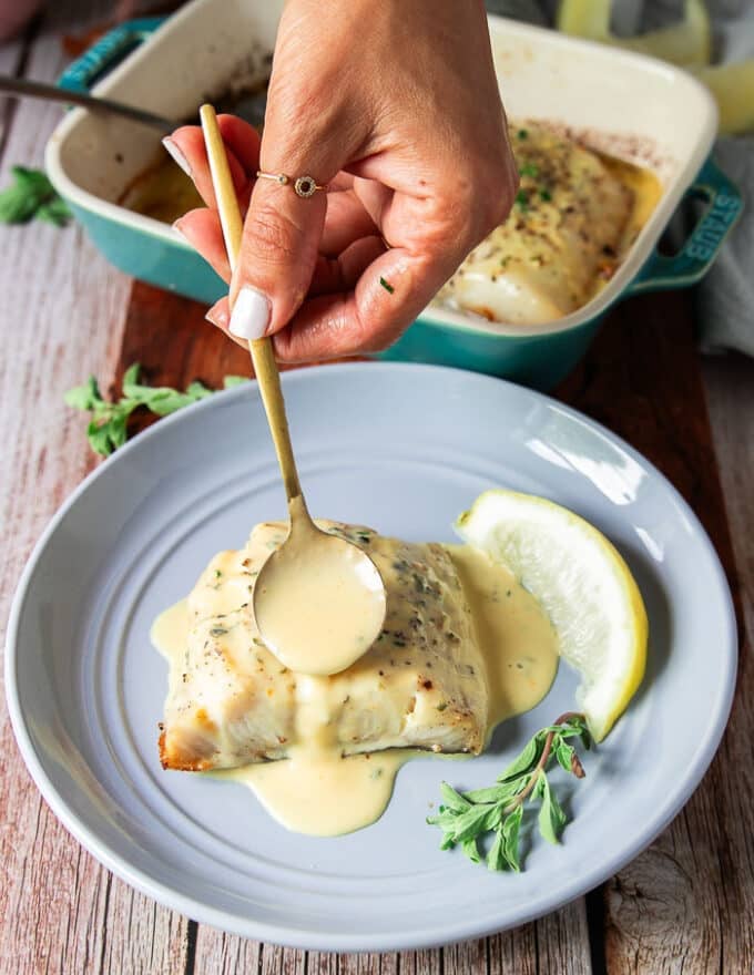 A hand spooning extra beurre blanc over the baked Chilean sea bass