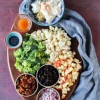 Ingredients to make broccoli cauliflower salad with raw broccoli florets, raw cauliflower florets, a bowl of cranberries , some copped onions, some crunchy wlanuts