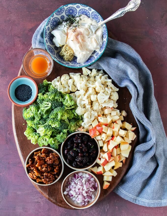 Ingredients to make broccoli cauliflower salad with raw broccoli florets, raw cauliflower florets, a bowl of cranberries , some copped onions, some crunchy wlanuts
