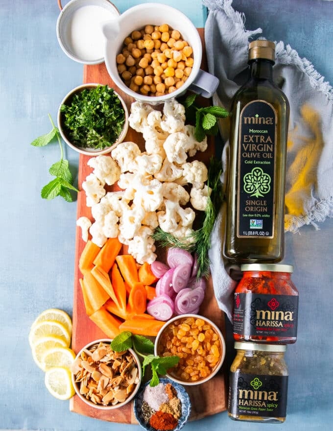 ingredients needed to make cauliflower salad including cauliflower florets, chickpeas, onions, carrots, arugula, raisins, almonds and a creamy tahini dressing, some warm spices in a bowl and harissa , and olive oil 