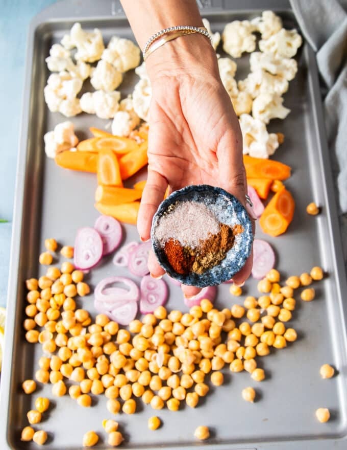 a hand holding a bowl of warm spices to season the cauliflower and chickpeas, onion and carrots before roasting 