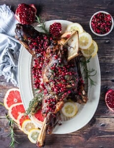a whole lamb shoulder roasted on a plate with pomegranate arils and rosemary and orange