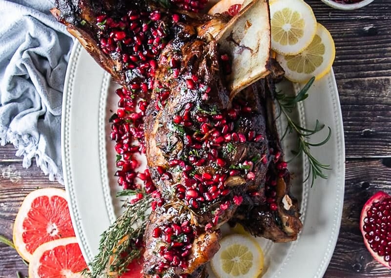 a whole lamb shoulder roasted on a plate with pomegranate arils and rosemary and orange