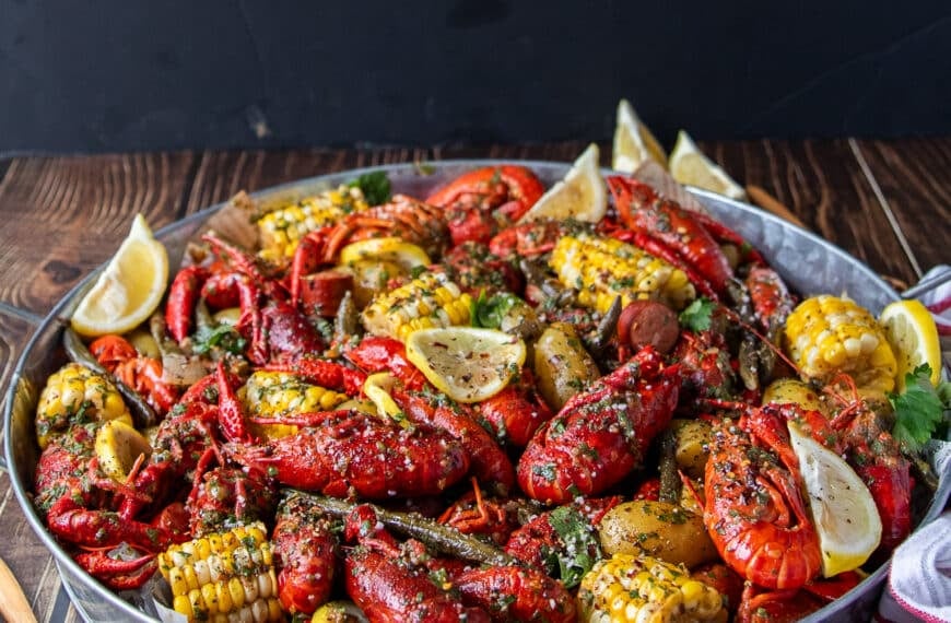 close up of a crawfish boil with crawfish , corn, potatoes, sausage, green beans and lemon wedges on a large platter ined with newspaper