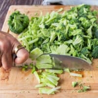 A hand holding a knife to thinly slice the broccoli stalks to make broccoli slaw