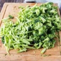 Broccoli stalks and florets are shredded thinly on a wooden board ready to make broccoli slaw