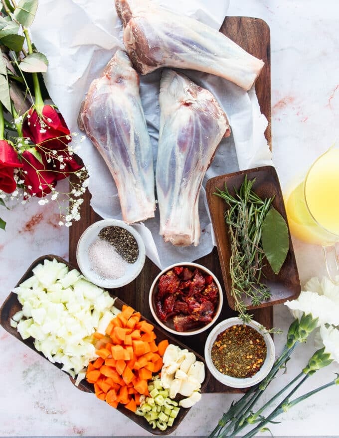 Ingredients needed to make slow cooked lamb shanks on a board with lamb shanks, small bowls of chopped aromatic, a plate of fresh herbs, some stock in a jar, some tomatoes and seasoning in a small plate 