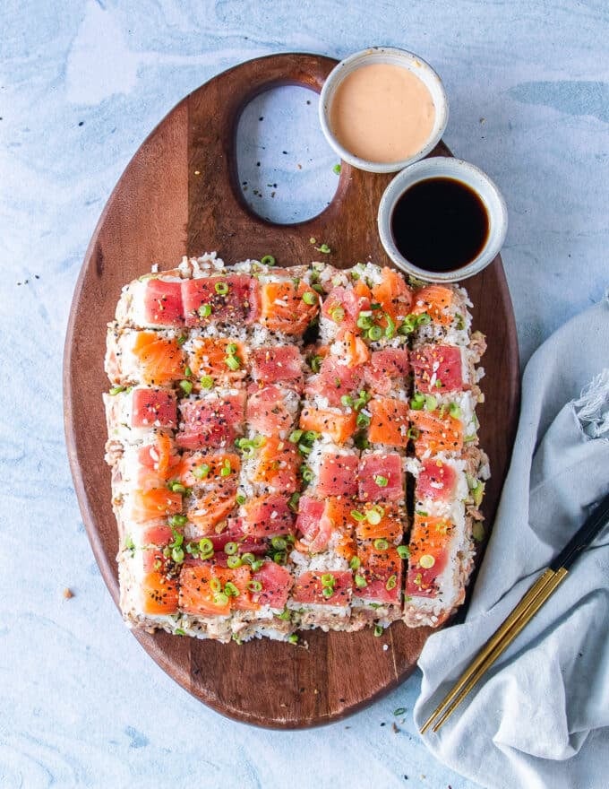 Top view of a square sushi cake on a wooden board topped with furikake, sesame seeds and green onions for garnish. A bowl of spicy mayo and soy sauce on the side for dipping.