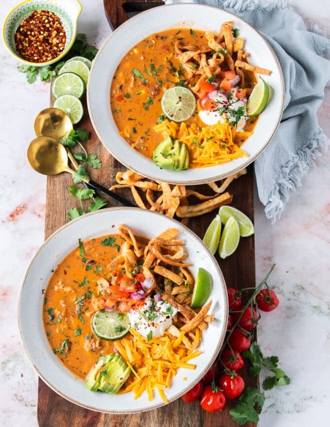 two bowls of chicken taco soup topped with tortilla chips, avocados, sour cream, and lime wedges 
