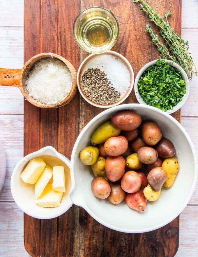 Ingredients to make smashed potatoes in a large board including the potatoes, butter, herbs, water, salt, seasoning, parmesan cheese 
