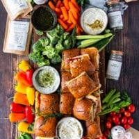 A large board with roast beef sliders with a variety of bowls of dips, some mini broccoli, mini peppers, mini carrots and a variety of veggies