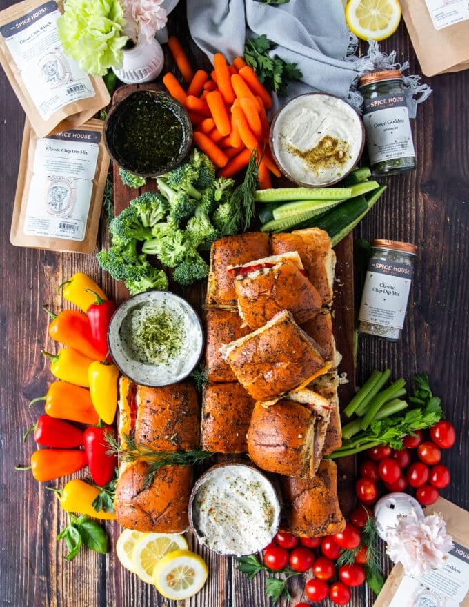 A large board with roast beef sliders with a variety of bowls of dips, some mini broccoli, mini peppers, mini carrots and a variety of veggies
