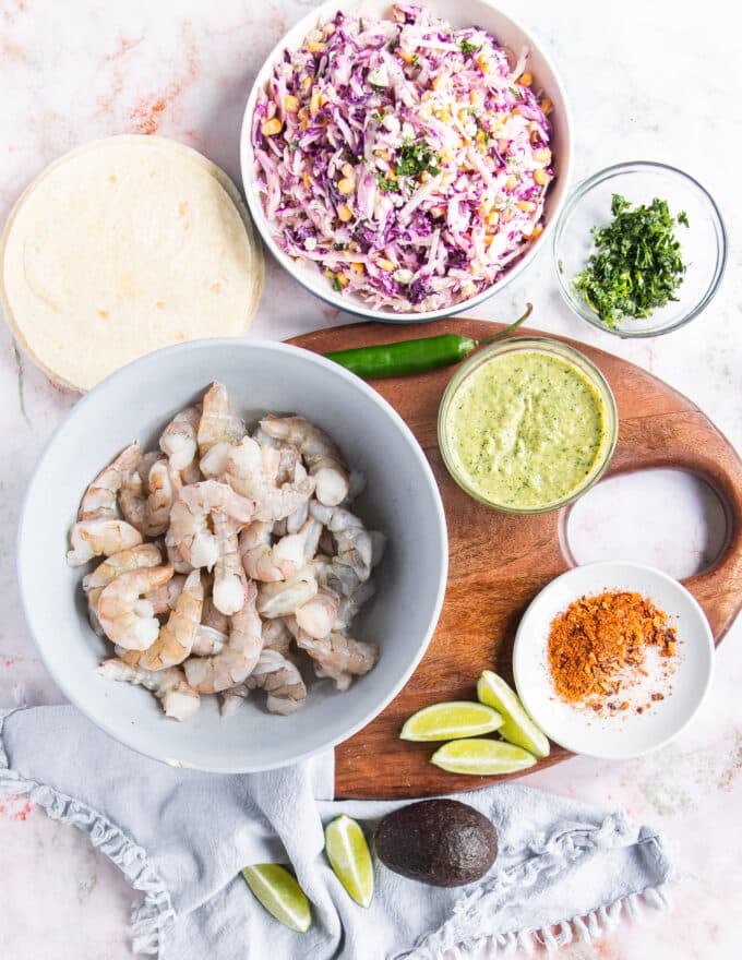ingredients for the shrimp tacos, including raw shrimp, taco seasoning, the tortillas, the poblano sauce, the corn cabbage slaw, the avocado, jalapeno, cilantro, lime and other toppings in small bowl