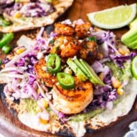 A close up of shrimp tacos assembled showing charred tortilla, creamy taco sauce, crunchy slaw, avocado slices, cilantro, cotija and lime