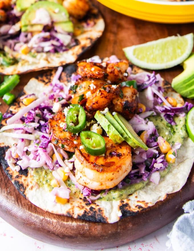 A close up of shrimp tacos assembled showing charred tortilla, creamy taco sauce, crunchy slaw, avocado slices, cilantro, cotija and lime
