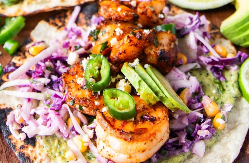 A close up of shrimp tacos assembled showing charred tortilla, creamy taco sauce, crunchy slaw, avocado slices, cilantro, cotija and lime