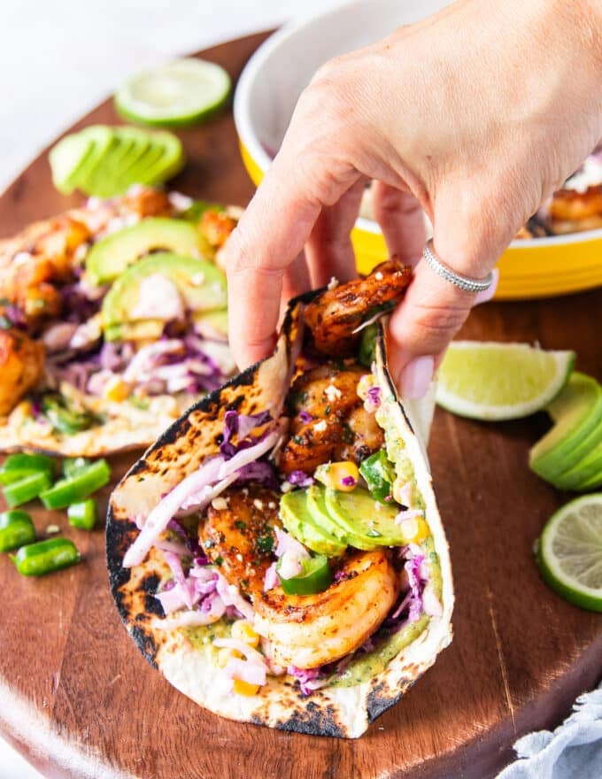 A hand taking a shrimp taco off the board showing the layered juicy shrimp and taco fillings