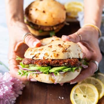 Two hands holding a tuna burger close up showing the layers of avocado, bun, tuna burger, lettuce and other toppings