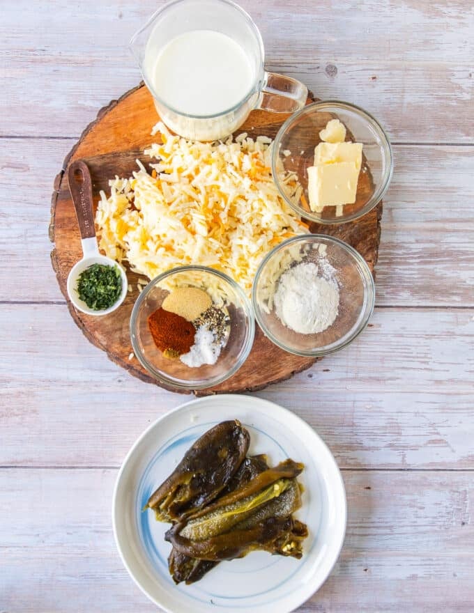 ingredients to make smoked queso on a board including cheeses, seasoning, herbs, roasted poblanos, milk, butter and flour