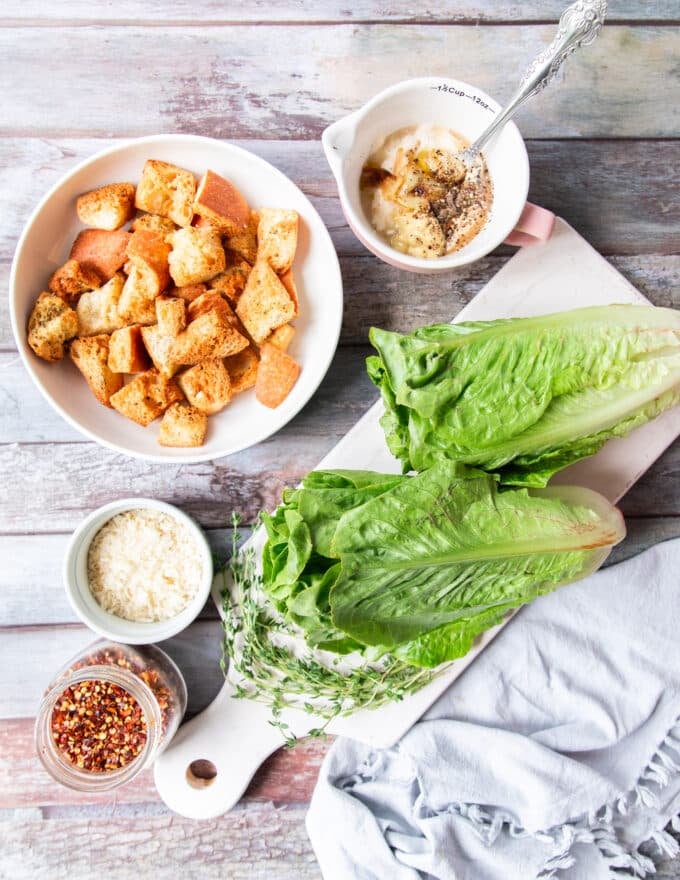 A board of all the ingredients to make grilled romaine salad including the romaine lettuce, a salad dressing ingredients , some seasoning, some parmesan cheese, croutons and toppings