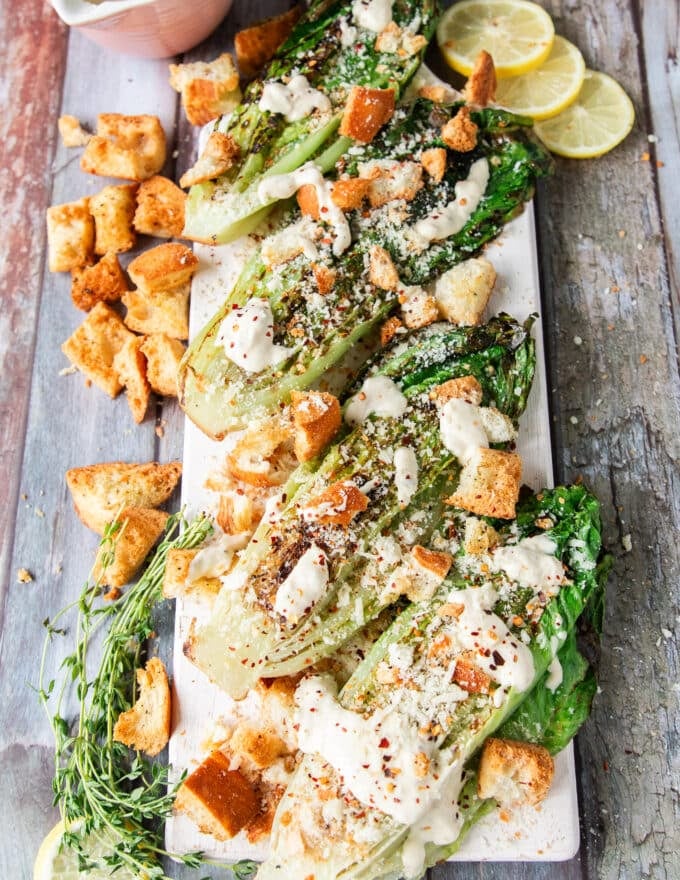 A fully dressed plate of several grilled romaine dressed up and topped with crunchy croutons and parmesan cheese and lemon wedges on the side.