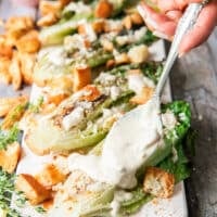A hand drizzling some creamy caesar dressing over the grilled romaine over a serving board
