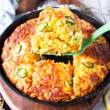 A spatula serving up a spoonful of jalapeno cheddar cornbread from the cast iron skillet showing the texture and studded corn and jalapeรฑo slices on top with a crisp cheese top