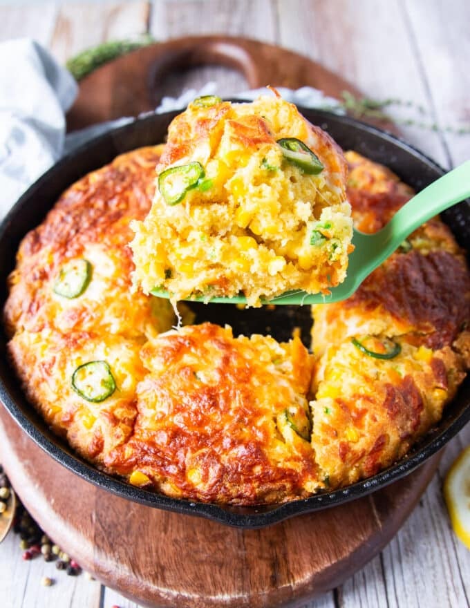 A spatula serving up a spoonful of jalapeno cheddar cornbread from the cast iron skillet showing the texture and studded corn and jalapeรฑo slices on top with a crisp cheese top