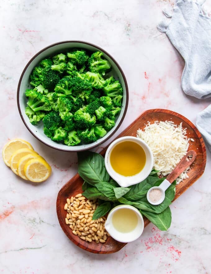 Broccoli pesto ingredients on a marble board with broccoli, parmesan, olive oil, garlic, lemon juice, basil, pine nuts and seasoning