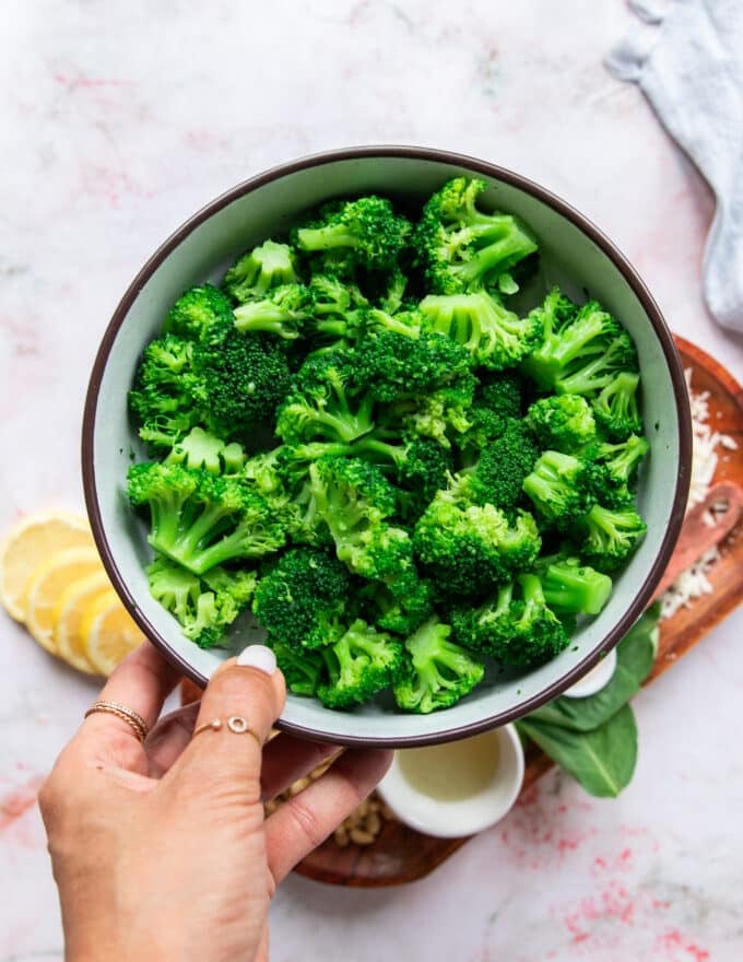 A hand holding a bowl of broccoli, blanched or frozen to be used to make the pesto
