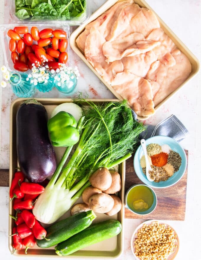 ingredients to make the chicken salad including chicken breasts, a variety of veggies, a bowl of seasoning blend, some greens, tomatoes and dressing ingredients in a bowl